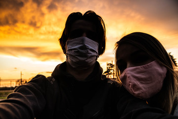 Dramatic portrait selfie of young couple with cotton and medical face masks with orange intense cloudy sunset in the background. Hard quarantine times concept. Apocalyptic. Emotional mood