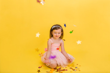 Obraz na płótnie Canvas A beautiful little girl in a puffy pink dress plays with confetti on a yellow isolated background with space for text. Birthday party
