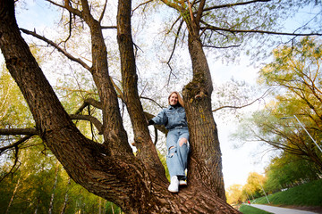 Joyful red-haired girl teenager in jeans climbed a branchy tree and laughs