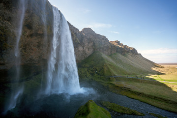 Seljalandsfoss during sunny day with lots of tourists approaching to walk behind the waterfall.