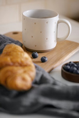 Morning breakfast or a snack with a ceramic cup of hot tea covered with stars and a croissant with blueberries. Kitchen towel and wooden board.