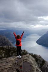 Trip to Norway. Brown hair woman in red jacket and black jeans feels freedom on the Preikestolen mountain (Preacher's Pulpit or Pulpit Rock) with Lysefjord on background in cloudy summer day