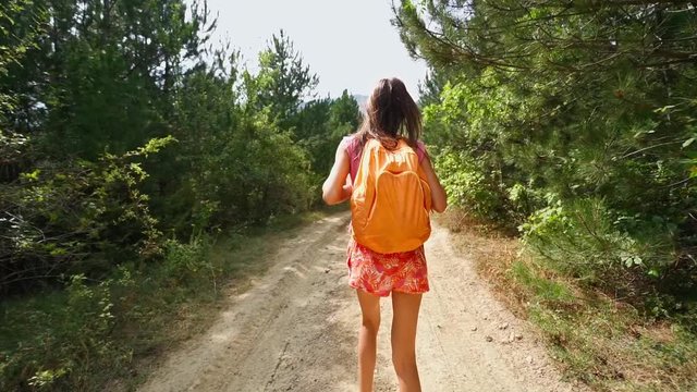 POV rear view happy smiling woman Backpacker with orange backpack walking along rough ground road with woods On sides. female hiker at sunny summer day enjoying live nature. super slow motion 120 fps