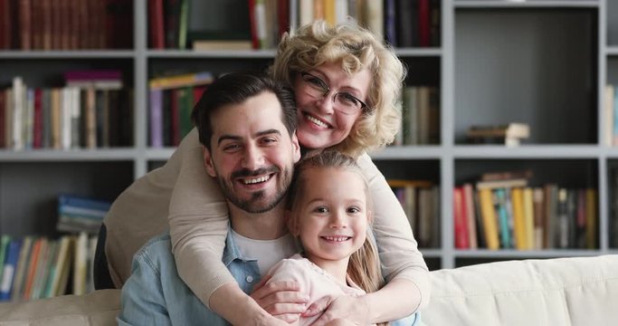 Head shot close up portrait excited middle aged beautiful woman embracing grownup smiling son and little child granddaughter at home. Happy three generations family cuddling, enjoying tender moment.