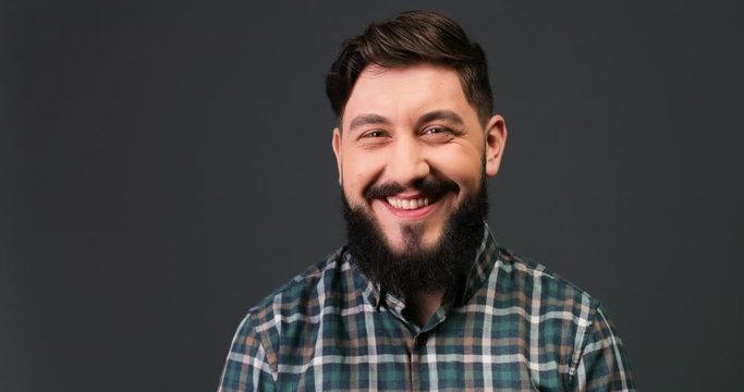 Close up portrait of handsome happy man with beard and mustache in checkered shirt smiling and winking to camera while standing on grey background indoor. Joyful funny Caucasian guy in good mood.