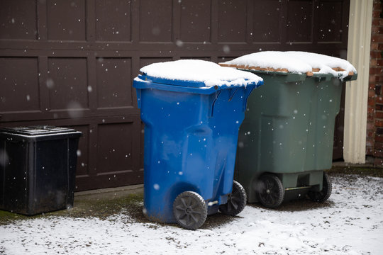 Snow covered recycle and garbage cans in front of a home garage