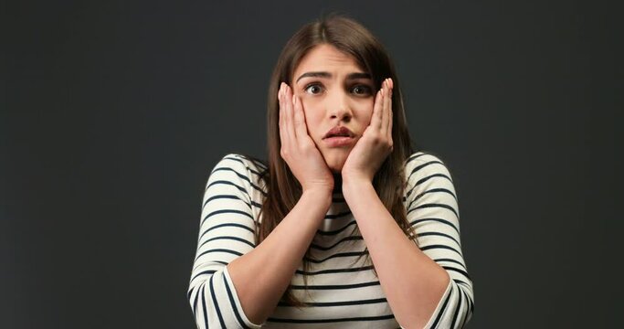 Close up portrait of scared disappointed Caucasian pretty girl with surprised face on dark-grey background. Beautiful young dark-haired woman with shocked face posing indoors. Emotions concept