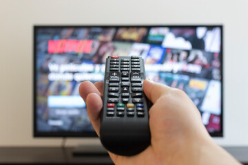 Closeup to a male hand holding a black TV controler with a blurred flat tv at background. indoor...