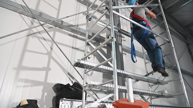 Caucasian Contractor Worker in His 30s Wearing Safety Harness Going Up Using Scaffolding To Replace Air Vent Elements Inside Warehouse Building.