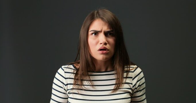 Close up portrait of scared Caucasian pretty girl with surprised face on dark-grey background. Beautiful young dark-haired confused woman looking away with shocked face posing indoors Emotions concept