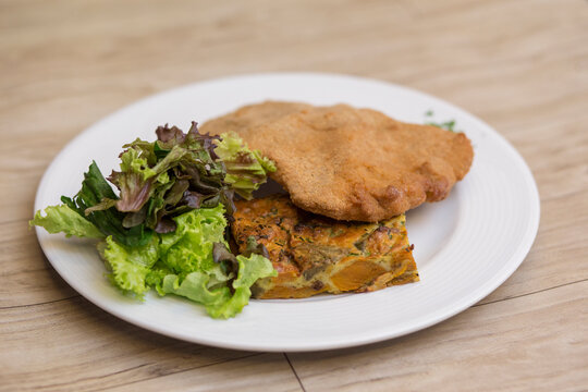 Milanesa, tarta de zapallito y lechuga en plato blanco redondo sobre mesa de madera