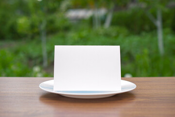 Blank standing bifold card on a classic empty white plate on the outdoor wooden table and green garden behind, clean template for you design presentation.