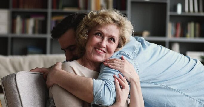 Excited blonde middle aged senior mother glad to see handsome grownup son at home. Smiling young man visiting mature elderly mommy, enjoying spending free leisure holiday time together indoors.