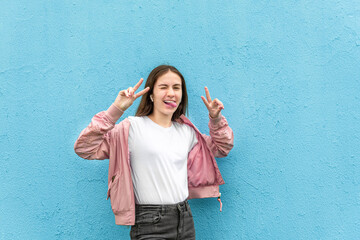 Portrait of funny caucasian teen girl in blank white t-shirt and pastel pink bomber jacket against blue city wall solid background. Emotion, generation Z, fashion, modern urban style, mockup concept.