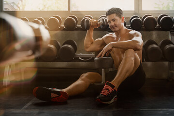 caucasian bodybuilder athletic man sitting alone on floor posing lifting dumbell with bottle of energy drink and dumbells aside during resting and relax after excercise and workout in gym and fitness