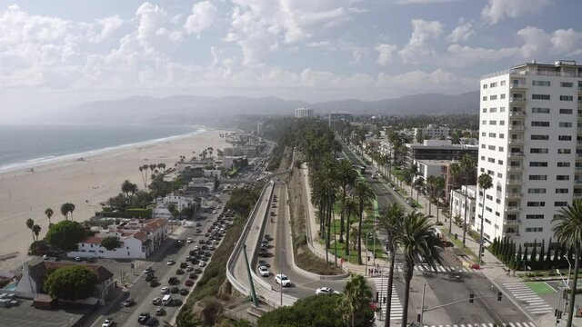 Aerial shot of street with palm trees by buildings and beach, drone flying forward in city against sky on sunny day - Santa Monica, California
