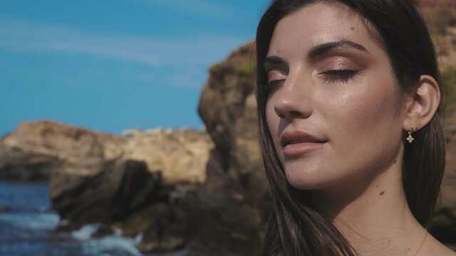 Short shot oa a model in a white dress walks down a cove of dark stones with the sea at her side hitting large rocks.