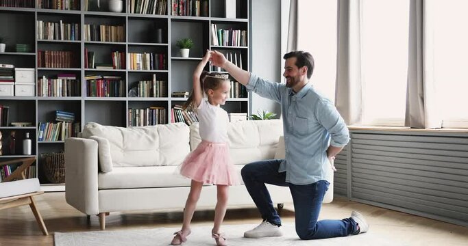 Full length smiling young daddy standing on one knee, twisting happy little preschool princess kid daughter, kissing hand after finishing dancing in modern living room, family hobby weekend pastime.