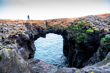 Obraz premium A tourist walks over a stone volcanic stone archway in Arnarstapi, West Iceland