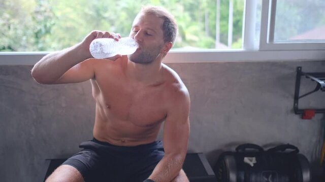 Muscular sport man drink water from bottle near window in gym with day light and he feel fresh and happy after drink. Concept of exercise or workout to get good health for prevention disease or covid.