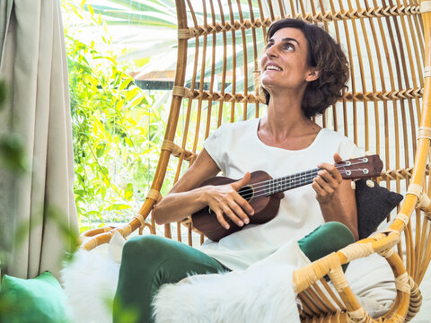 happy and relaxed woman playing ukulele sitting in a bamboo rattan hanging swing chair with green plants