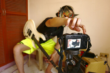 Asian teenager with a character mask filming himself while playing an electric guitar in his room