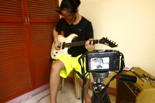 Asian teenager with a character mask filming himself while playing an electric guitar in his room