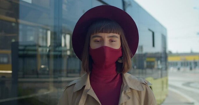 Portrait of millennial girl in protective cotton mask turning head and looking to camera.Close up view of young woman standing outdoors near public transport. Concept of virus protection.