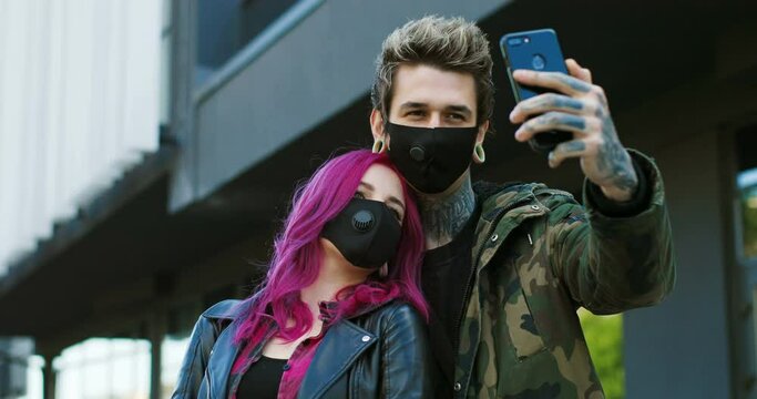 Caucasian extraordinary couple of hipsters in respiratory masks standing at street and taking selfie photos with smartphone camera. Stylish girl with pink hair and guy outdoor making photo with phone.