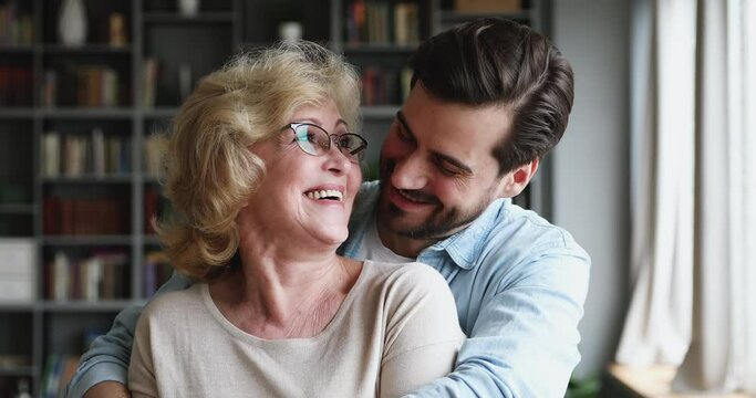 Close up portrait happy millennial bearded man cuddling beautiful senior elderly mother. Head shot affectionate grownup son showing love and care to smiling middle aged mature mommy in living room.