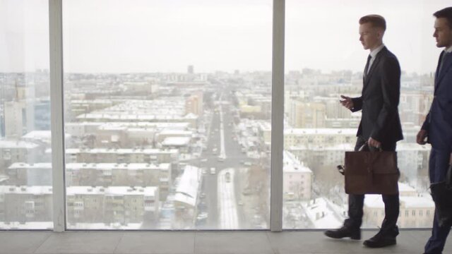 Medium shot with slowmo of businessmen in suits carrying briefcases and walking before panoramic window while discussing work. Businesswoman in formalwear walking past camera