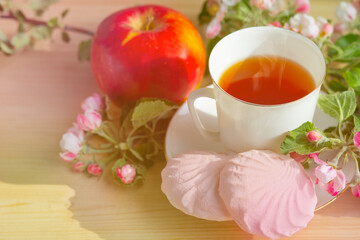 On a wooden background, a branch of apple flowers, a mug with tea, marshmallows and red apples. Still life. Close-up, food and spring concept, postcard. Copy space, space for text.