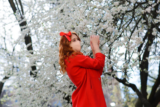 Close up portrait of tender red-haired girl in a red coat under a blossoming cherry tree with a mask with flowers on from the coronavirus.