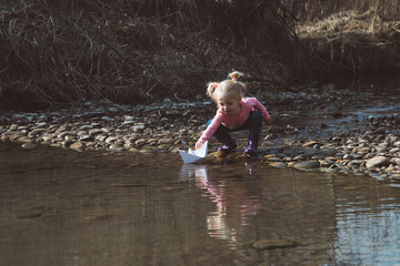 Little girl in rubber boots launches paper white boats in creek in spring or autumn