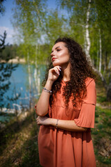 A girl with long wavy curly hair in her hair woven flowers in a white transparent guipure dress barefoot in the summer on nature in a forest at sunset standing standing near the road and bushes