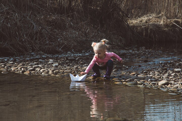 Little girl in rubber boots launches paper white boats in creek in spring or autumn