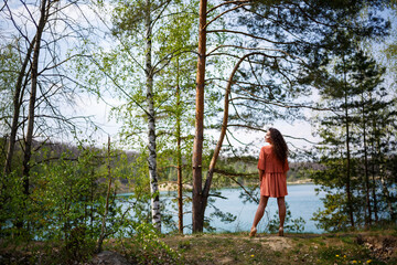 Wood masonry. A girl with long wavy curly hair in an orange guipure dress and shoes on nature, in a forest by the lake, stood near trees and bushes. Young woman smiles and enjoys life