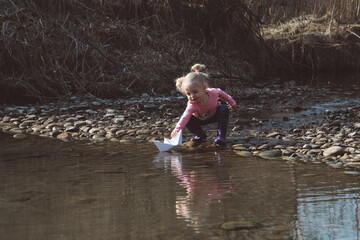 Little girl in rubber boots launches paper white boats in creek in spring or autumn