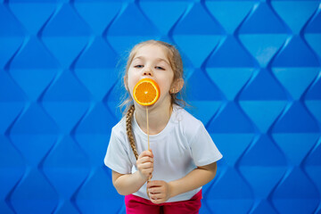 little happy girl with an orange in her hand, looks at the camera dressed in White T-shirt, isolated on blue background, copy space,