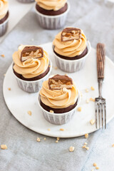 Peanut butter cupcakes with chocolate bites and chopped nuts on grey linen tablecloth.