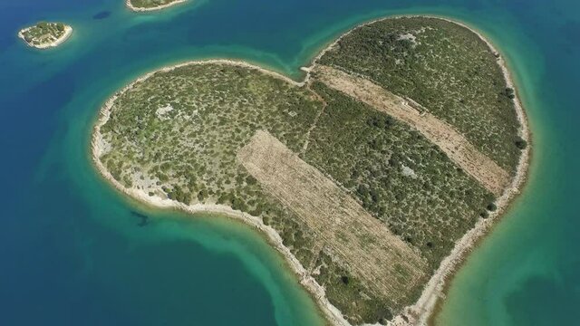 Aerial panning shot of small heart shape island in ocean on sunny day, drone flying backward over sea - Biograd, Croatia