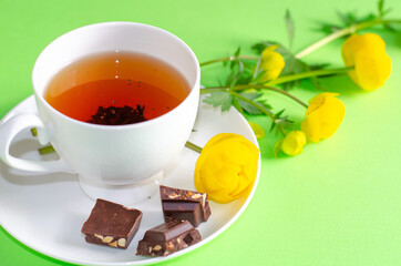 A Cup of black tea, pieces of chocolate and yellow flowers of the Trollius europaeus on a green background. Sunny, bright, shadows fall from the window. Chocolate day and morning tea ceremony.