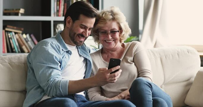 Happy young bearded man showing kids funny photos on smartphone to smiling middle aged senior mother in eyeglasses. Positive grownup millennial son teaching smart older mature mommy using mobile apps.