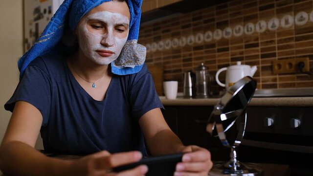 Young sad asian woman works in the kitchen. Self-isolation. Girl in a clay mask and with a towel on her head using her smartphone for social media, communications or texting, read news