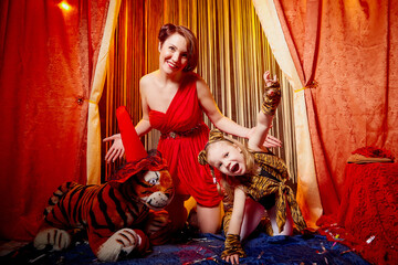 Fototapeta na wymiar Family during a stylized theatrical circus photoshoot in a beautiful red location. Models mother who looks like a trainer and daughter who looks like a tiger pose on stage with curtains