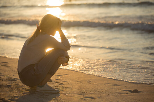Sad depressed young woman sitting alone with hand on face at the beach near the water during sunset. Silhouette 