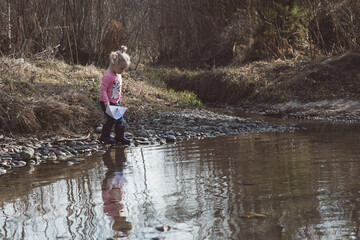 Little girl in rubber boots launches paper white boats in creek in spring or autumn