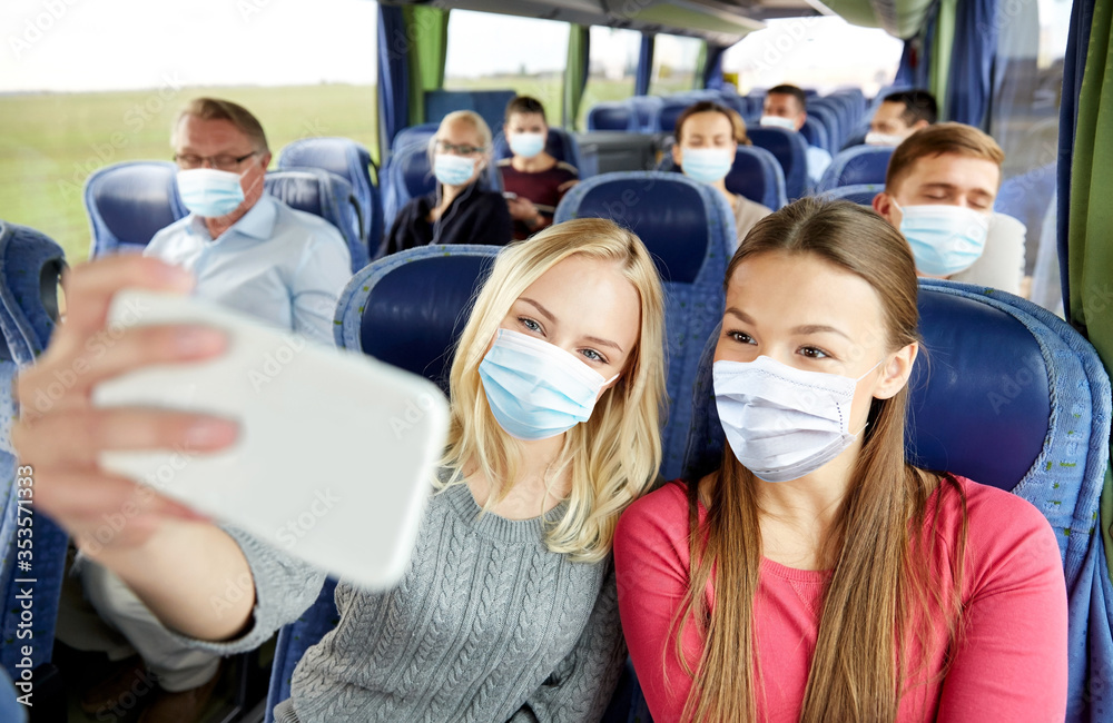 Poster transport, tourism, road trip and people concept - young women or friends wearing face protective medical mask for protection from virus disease in travel bus taking selfie by smartphone