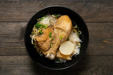 Rice noodle with chicken leg in black bowl on wooden table.