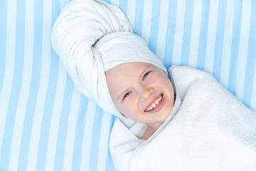 Fototapeta na wymiar Portrait of cute little caucasian girl wrapped in white towel with bath towel on head over striped blue bed background. Spa procedure, bath textile.